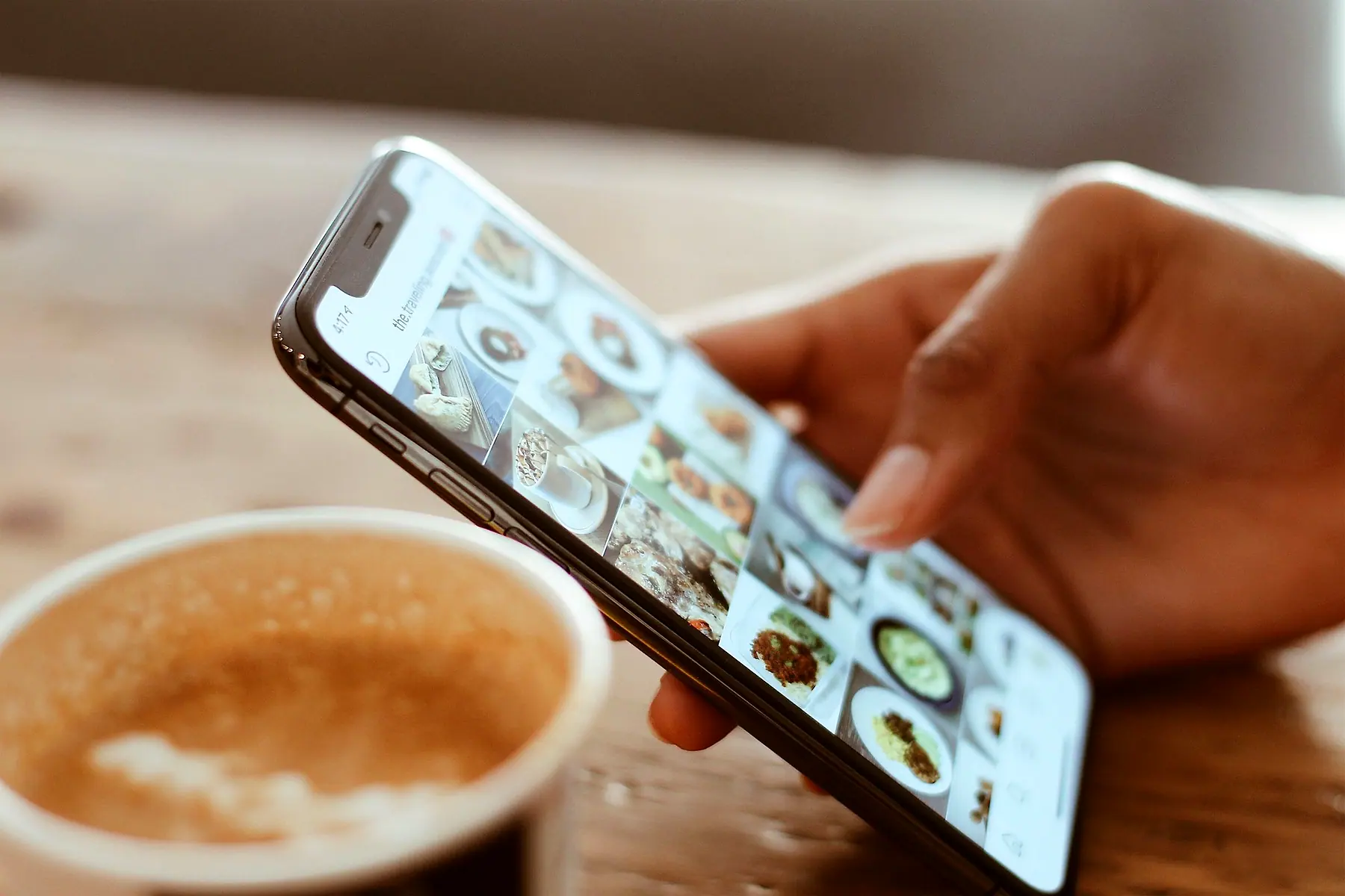 A hand scrolling a feed of photos on a phone beside a cup of coffee