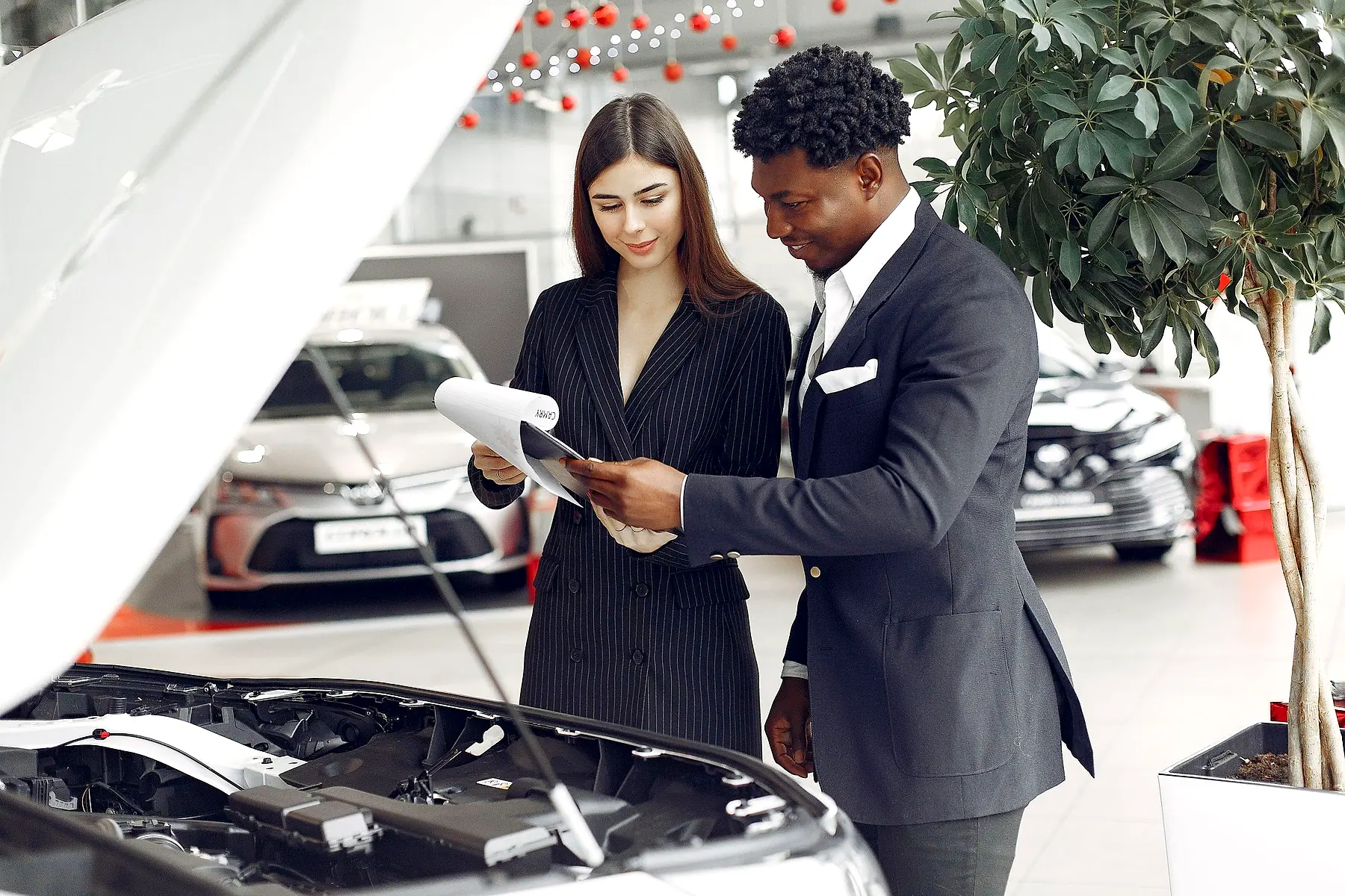 A salesperson and a customer reviewing a clipboard beside a car with its bonnet open in a showroom