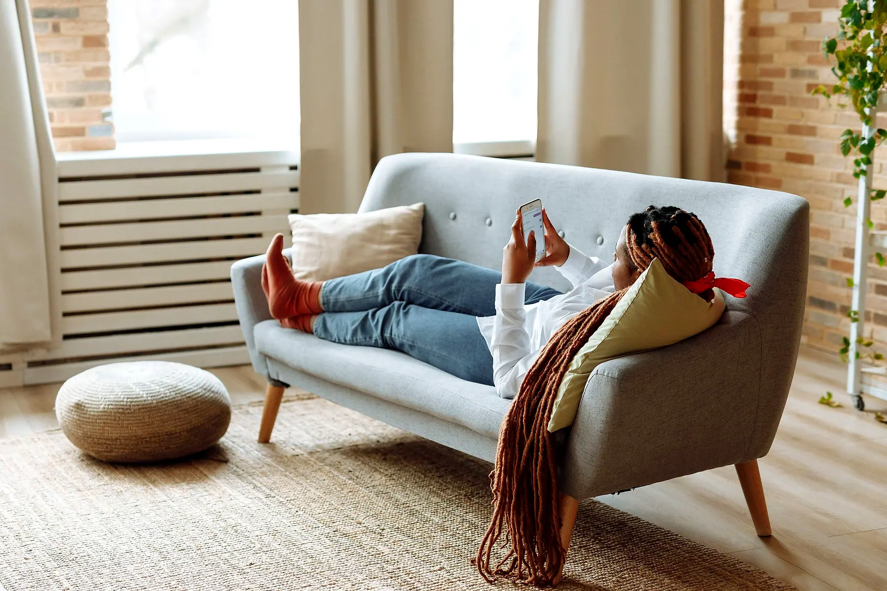 A woman with long braids lying on a grey sofa looking at her phone